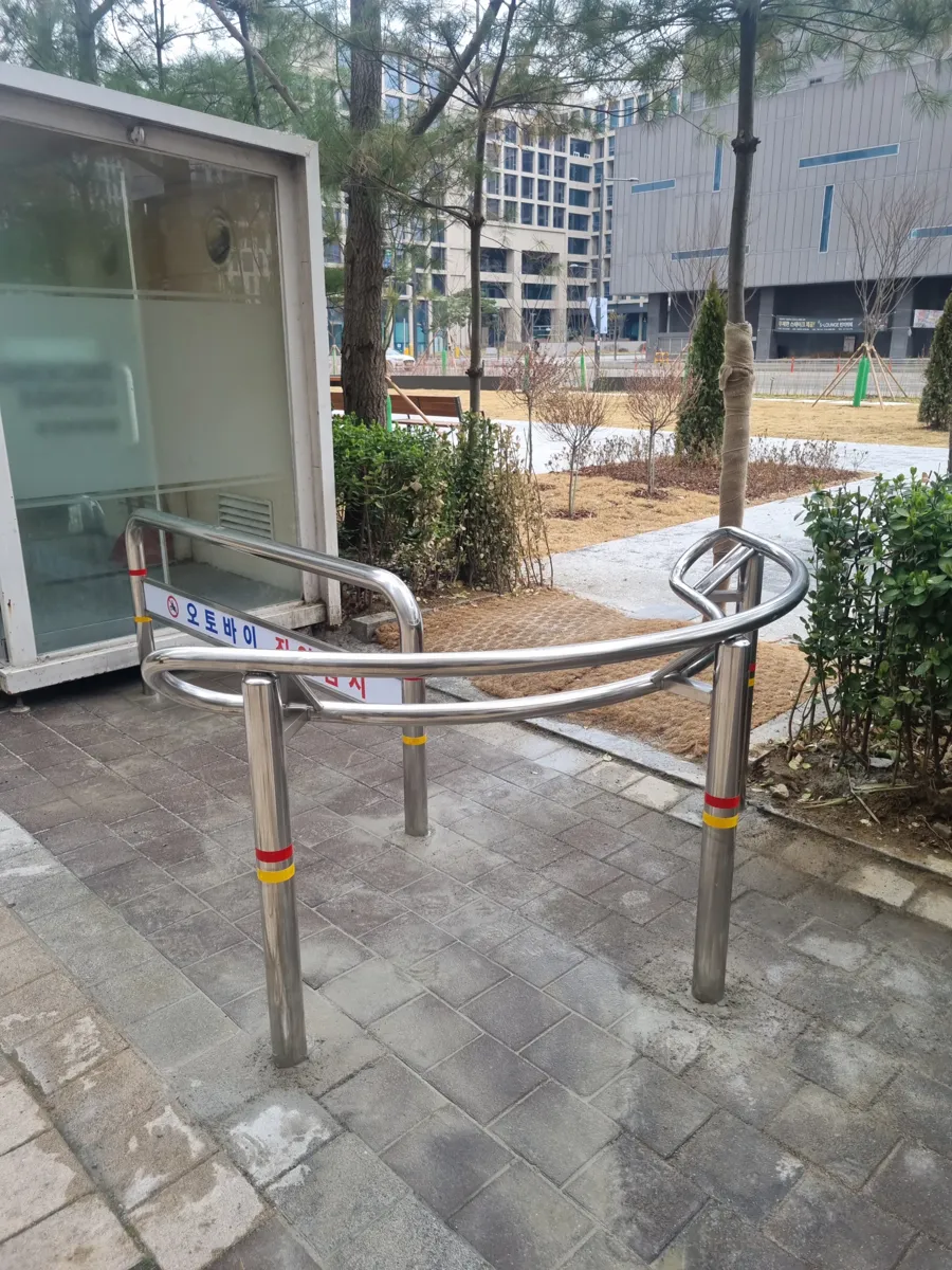 유리 부스 옆 수목이 있는 보행로 모서리에 설치된 곡선 레일형 스테인리스 볼라드 근접 모습